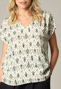 Blouse blanche avec un décolleté en V, à manches courtes, arborant un motif géométrique vert et noir avec des motifs complexes tout au long.