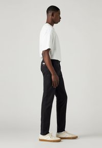 Black tapered pants with a patch pocket and logo detail, paired with a white short-sleeve T-shirt and beige sneakers. Smooth fabric texture.