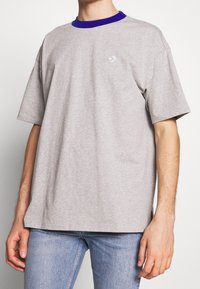 Hombre vistiendo una camiseta oversized gris claro con un cuello azul y un pequeño logo blanco en el pecho, combinada con jeans azul claro.