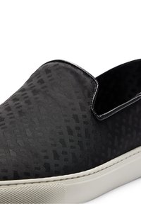 Baskets noires à enfiler avec un motif texturé subtil et une semelle en caoutchouc blanche, montrant les détails latéraux et supérieurs sur un fond blanc.