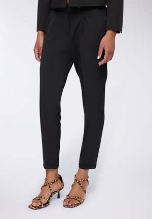 Pantaloni neri su misura con pieghe e orli ripiegati, abbinati a sandali con tacco alto a strisce in stampa leopardata, dotati di sottili cinghie alla caviglia.