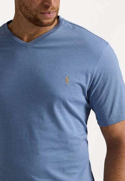 Světle modré v-neck tričko vyrobené z hladké bavlněné směsi, s malým vyšitým zlatým logem na levé straně hrudi.