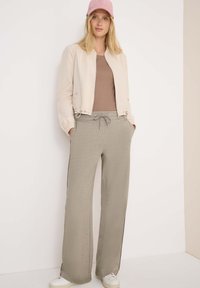 Veste bomber beige clair, haut ajusté marron, joggings gris à jambes larges avec cordon de serrage. Une casquette rose et des baskets blanches complètent le look.