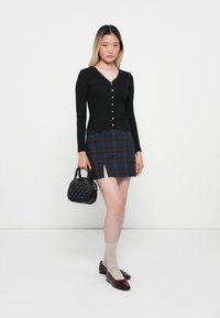 Cardigan noir à côtes avec boutons en perles, superposé sur une jupe courte à carreaux dans des tons de bleu et de marron. Sac à main tressé noir et chaussettes beige.