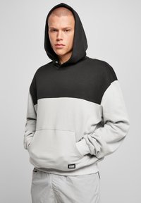 Giovane uomo con una felpa nera e grigia con cappuccio, con le mani nella tasca frontale, in piedi contro uno sfondo grigio chiaro uniforme.