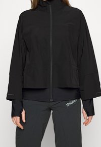 Svart jacka med hög krage, dragkedja och vida ärmar. Har en slät textur och subtila utsmyckningar nära ärmsluten.