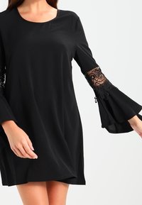Svart klänning med långa klockärmar försedda med intrikata spetsmönster. Slät textur med rund halsringning och lös passform.