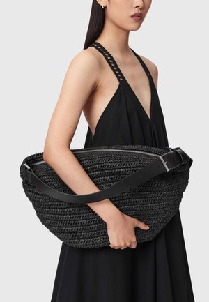 Γυναίκα με μαύρο αμάνικο φόρεμα κρατά μεγάλο πλεκτό μαύρο τσαντάκι με φαρδιά δερμάτινη λαβή και κλείσιμο με φερμουάρ.