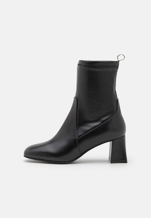 Bottine noire avec une finition lisse, talon bloc, bout arrondi, détails de surpiqûres latérales et languette à l’arrière, présentée sur fond blanc.