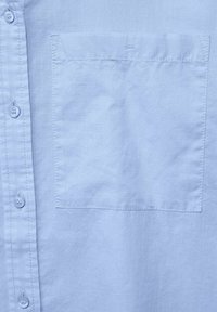 Světle modrá košile na knoflíky s hladkým povrchem a jednou kapsou na hrudi. Obsahuje osm knoflíků na léze a jemné prošívání.