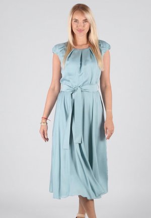 Frau mit langen blonden Haaren, die ein hellblaues, tailliertes Midikleid trägt und vor einem schlichten grauen Hintergrund steht.