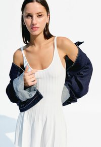 Biele rebrované tank šaty s rozšíreným lemom, vrstvené pod oversized tmavomodrou džínovou bundou so šedými rukávmi. Jednoduchý a štruktúrovaný dizajn.