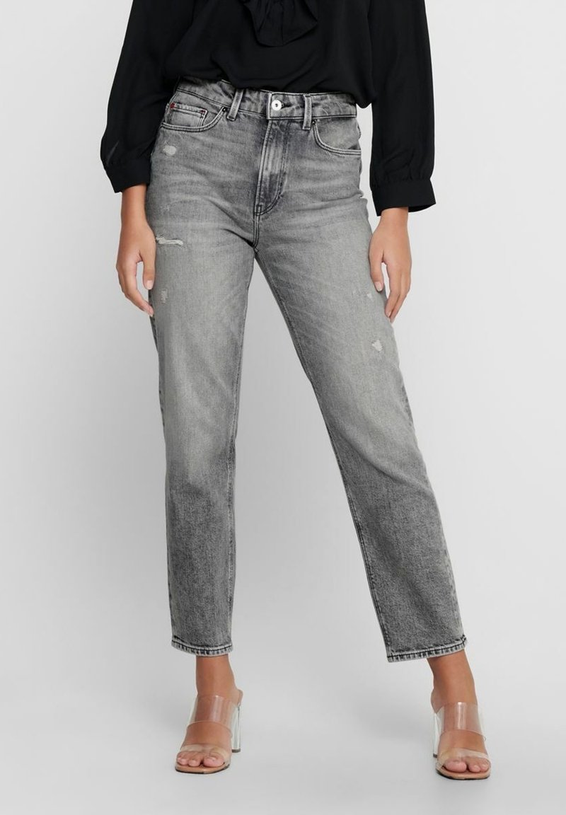 Femme portant un jean gris délavé taille haute légèrement usé, une blouse noire à manches longues et des sandales à talons transparents.