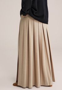 Vista laterale di una persona che indossa una maxi gonna plissettata beige, un maglione nero e stivali alla caviglia marroni con le mani in tasca.
