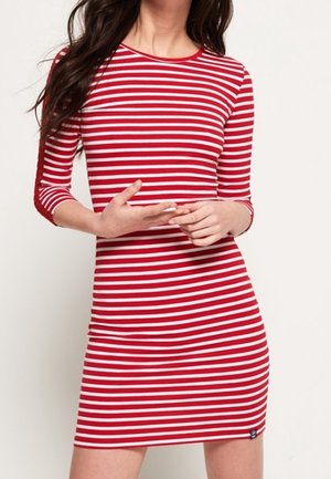 Kvinde iført en rød og hvid kjole med vandrette striber og trekvartlange ærmer, stående foran en ensartet lys baggrund.