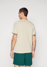 Beige kortærmet T-shirt med rund halsudskæring, glat tekstur og flad kant, båret sammen med teal shorts. Synlige tatoveringer på armene.