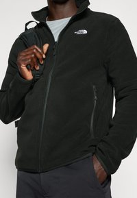 Mustat fleece-takki, jossa on korkea kaulus, vetoketjullinen etuosa ja vasemmassa rinnassa logo. Ominaisuuksiin kuuluu vetoketjullinen sivutasku ja pehmeä tekstuuri.