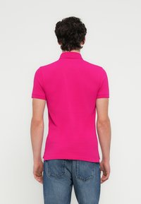 Εφαρμοστό πόλο μπλουζάκι με κοντά μανίκια σε έντονο ροζ ύφασμα, με δομημένο γιακά, ομαλή υφή και πλευρικές σχισμές, συνδυασμένο με μπλε τζιν παντελόνι.