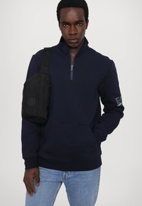 Sudadera de cremallera azul marino con un cuello alto, que cuenta con un bolsillo frontal y puños acanalados. El modelo lleva un bolso cruzado negro.
