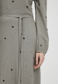 Grau gefärbtes Kleid aus Stoff mit schwarzen Pünktchen, lange Ärmel, geraffte Manschetten und ein gebundenes Taillendetail. Glatte Textur.