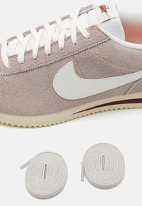 Nike sneaker világosszürke velúrbőr felsőrésszel, fehér bőr Swoosh-al, krémszínű talppal és texturált talppal; két kerek világosszürke fűzős szalagot tartalmaz.