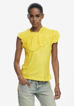 Yellow short-sleeve top with embroidered swirl patterns, textured frayed edges, and a relaxed fit. Paired with light blue jeans.