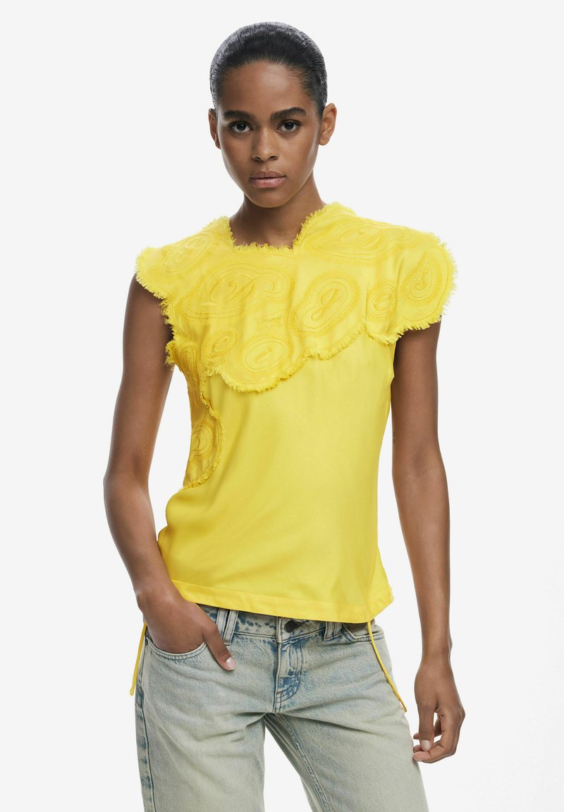 Gelbes T-Shirt mit kurzen Ärmeln, bestickten Wirbelmustern, strukturierten, ausgefransten Kanten und einem lässigen Schnitt. Kombiniert mit hellblauen Jeans.