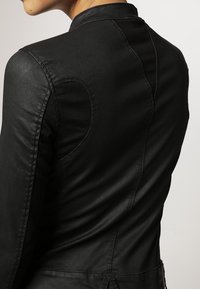 Vue de dos d'une personne portant une veste en cuir noire ajustée avec des coutures apparentes et des détails de surpiqûres sur les épaules et le dos.