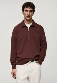 Burgundská mikina na zip s límcem, vyrobená z měkkého materiálu. Má dlouhé rukávy a úzký zip u krku. Doplněna bílými kalhotami.