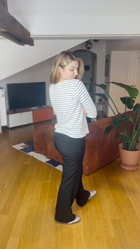 Baltas ir tamsiai mėlynas dryžuotas ilgomis rankovėmis viršus, juodos platėjančios kelnės, sidabrinės buto bateliai, vidaus interjeras su augalais ir matomu baldais.