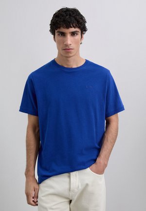 Joven con cabello rizado oscuro, vistiendo una camiseta azul de manga corta sin estampado y pantalones beige claros, de pie contra un fondo claro y liso.