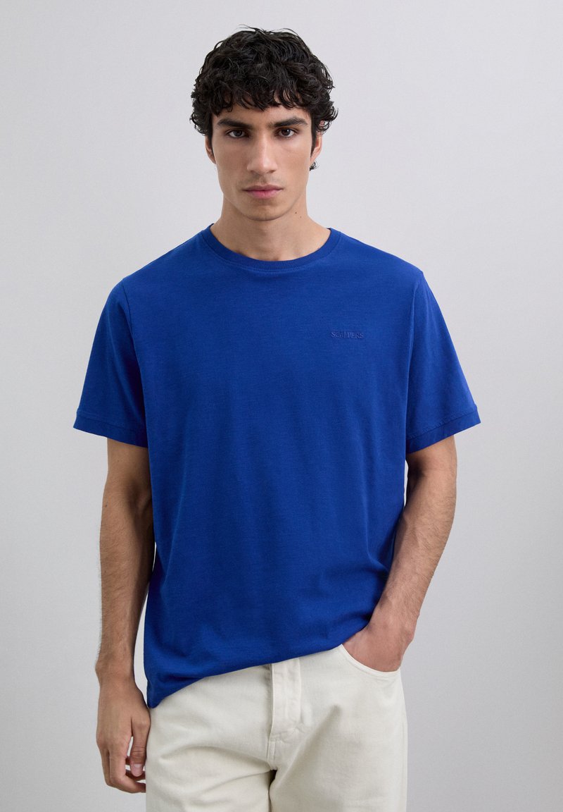 Joven con cabello rizado oscuro, vistiendo una camiseta azul de manga corta sin estampado y pantalones beige claros, de pie contra un fondo claro y liso.