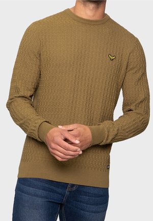 Homme portant un pull en maille marron texturé avec des poignets et un ourlet côtelés, associé à un jean bleu, les mains jointes devant lui.