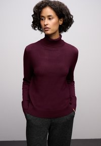 Burgundský rolák vyrobený z měkké látky. Design zahrnuje dlouhé rukávy a volný střih, doplněný o texturované šedé kalhoty.