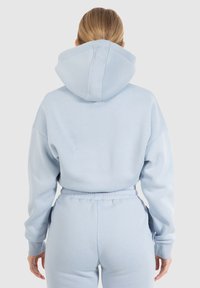 Felpa azzurra chiara con un ampio cappuccio, spalle scese e polsini a costine. Tessuto morbido; indossata con pantaloni abbinati, che mostrano un elastico in vita.