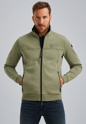 Veste à zip olive avec une partie supérieure à motif en losanges, poches latérales et poignets côtelés. Tissu doux avec une texture lisse.