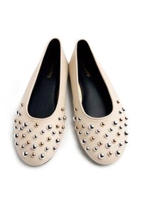 Zapatos planos beige hechos de cuero suave, con tachuelas metálicas en tonos plata y oro en la parte delantera, y un forro interior negro.
