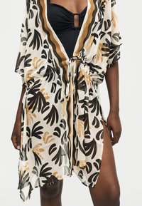 Maillot de bain noir avec centre froncé associé à un couvre-chef blanc transparent à manches courtes avec motifs abstraits de feuilles noires, brunes et beiges et taille à nouer.