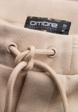Beige trekkingelastische tailleband met een gemêleerde stof, met een label gemarkeerd "ombre" en een geknoopt koord. Er zijn oogjes voor het trekkingelastiek.
