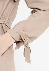 Beżowy, lekki materiał z rękawem z detalem w postaci zawiązanego supełka przy mankiecie. Posiada przeszycia oraz gładką fakturę.