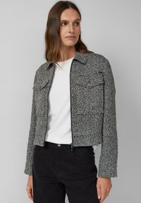 Kobieta ubrana w czarną i białą teksturowaną kurtkę zapinaną na zamek, nosi białą koszulę i czarne spodnie, patrzy w bok na tle jednolitego tła.