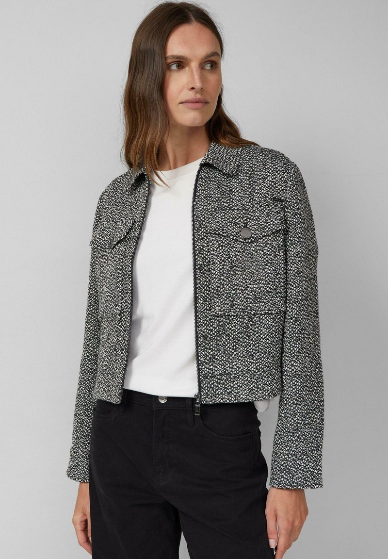 Kobieta ubrana w czarną i białą teksturowaną kurtkę zapinaną na zamek, nosi białą koszulę i czarne spodnie, patrzy w bok na tle jednolitego tła.