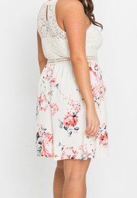 Květinové šaty s krajkovou tělovou částí v krémové barvě, páskem v oblasti pasu a rozšířenou sukní se růžovými a modrými květinami na světlém pozadí.