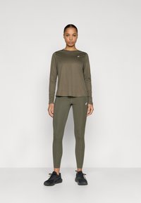 Μπλούζα μακριών μανικών σε ελιά πράσινο και κολάν από ελαφρύ, ελαστικό ύφασμα, συνδυασμένα με μαύρα αθλητικά παπούτσια. Λογότυπα διακοσμούν και τα δύο κομμάτια.