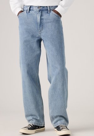 Person iført lyseblå løstsiddende jeans med hænderne i lommerne og sorte lave sneakers med hvide snørebånd på lys baggrund.
