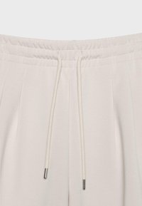 Pantalon de survêtement couleur crème avec une taille élastique et des cordons blancs munis d'embouts métalliques, montrés de face au niveau de la taille.