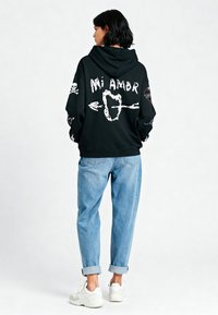 Felpa nera con grafica "Mi Amor" e varie illustrazioni bianche, abbinata a jeans azzurri arrotolati e sneakers bianche.