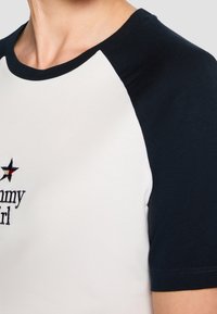 Biała koszulka z granatowymi rękawami raglanowymi, ozdobiona haftowanym napisem "Tommy Girl" i gwiazdkowym detalem na klatce piersiowej. Gładka tekstura.