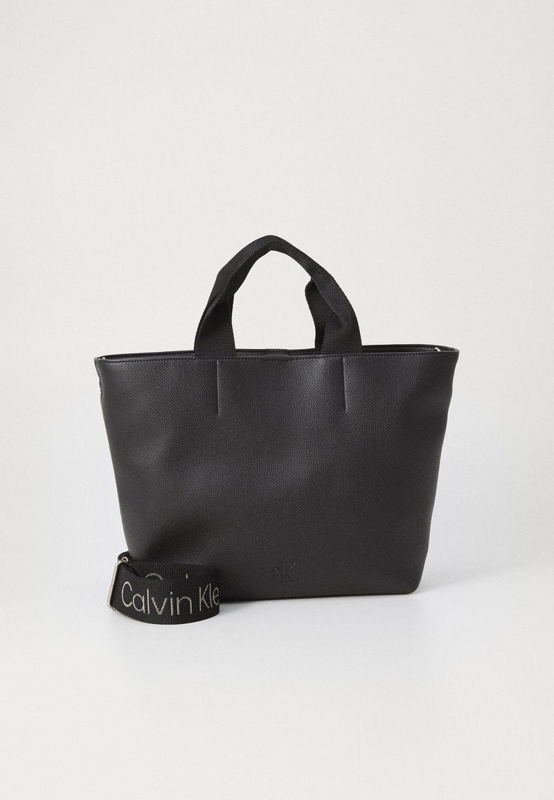Borsa tote nera realizzata in materiale testurizzato con due manici. Presenta una tracolla removibile con "Calvin Klein" in lettere bianche.