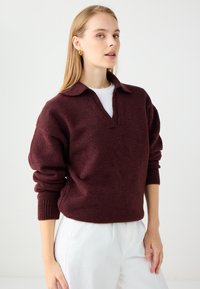 Burgundowa sweter wykonany na drutach z kołnierzykiem polo, w luźnym kroju, z prążkowanymi mankietami i gładką fakturą, zestawiony z białymi spodniami.
